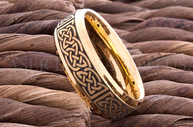 May include: A gold-colored ring with a black Celtic knot design. The ring is set against a background of brown rope. The ring's interior is a bright gold color, and the exterior features an intricate, repeating pattern.