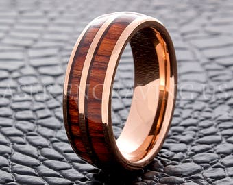May include: A rose gold ring with a wood inlay. The ring features a polished rose gold band with two parallel inlays of dark wood. The wood is a rich brown color, and the ring has a high-shine finish.