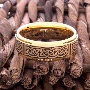 May include: A gold-colored ring with a black Celtic knot design. The ring is set against a textured, brown background. The ring's band is approximately 8mm wide.