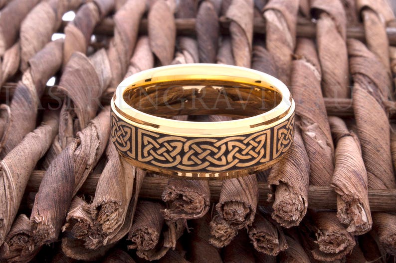 May include: A gold-colored ring with a black Celtic knot design. The ring is set against a textured, brown background. The ring's band is approximately 8mm wide.