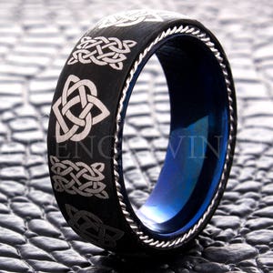 May include: A black and blue tungsten carbide ring with a silver Celtic knot design. The ring has a brushed finish and a beveled edge.