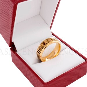May include: A gold-colored ring with a black Celtic knot design, displayed inside a red textured jewelry box with a white interior. The ring is the focal point, showcasing intricate details.