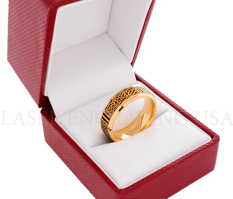 May include: A gold-colored ring with a black Celtic knot design, displayed inside a red textured jewelry box with a white interior. The ring is the focal point, showcasing intricate details.
