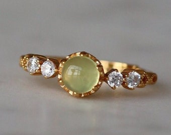 Puede incluir: Un anillo de oro con una piedra preciosa verde y pequeñas piedras preciosas transparentes. El anillo tiene un diseño delicado.