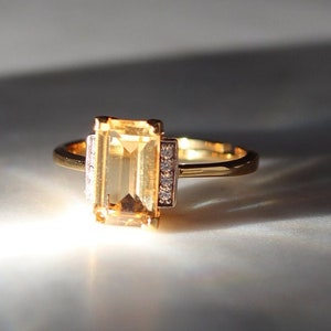 May include: A gold ring with a rectangular, pale-yellow gemstone. The ring has a slender band and small, clear accent stones on either side of the main stone. The ring is set against a soft, neutral background, with light and shadow effects.