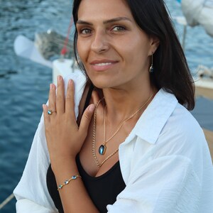 May include: A woman wearing a white shirt, black tank top, and gold jewelry with blue gemstone accents. She is wearing a ring, bracelet, necklace, and earrings. The jewelry features oval-shaped blue gemstones.