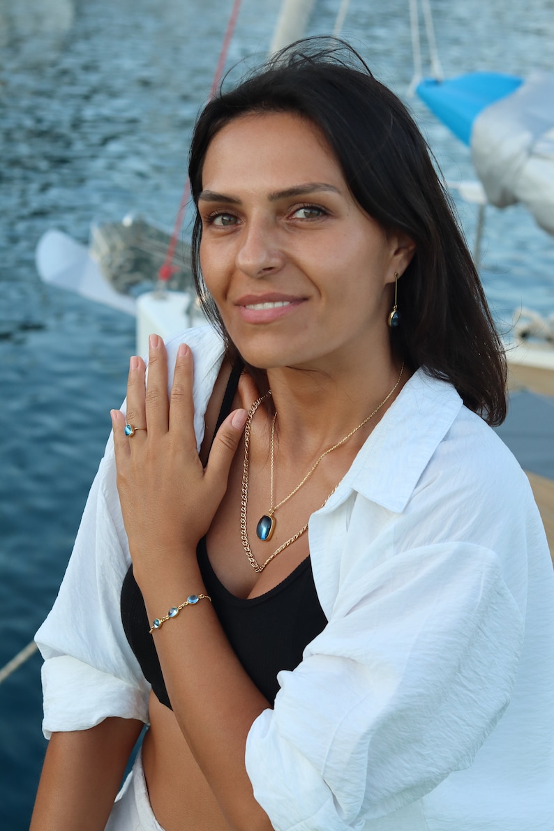 May include: A woman wearing a white shirt, black tank top, and gold jewelry with blue gemstone accents. She is wearing a ring, bracelet, necklace, and earrings. The jewelry features oval-shaped blue gemstones.