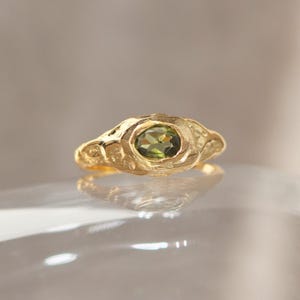 Peut inclure: Une bague en or ornée d'une pierre précieuse ovale vert olive. La bague présente un design texturé et organique. La pierre est sertie dans un sertissage à lunette, et le corps a un aspect sculpté unique. La bague est présentée sur une surface réfléchissante.