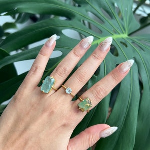 May include: Three gold rings with different green gemstone settings. The rings are on a hand with white nail polish and gold glitter accents.