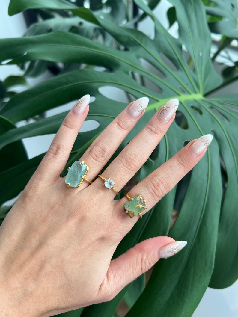 May include: Three gold rings with different green gemstone settings. The rings are on a hand with white nail polish and gold glitter accents.