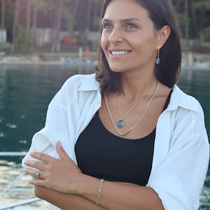May include: A woman wearing a white shirt, black tank top, and white pants. She is wearing a gold necklace with a blue gemstone pendant, a gold bracelet with blue gemstones, and a gold ring with a blue gemstone. She is smiling and looking to the right.
