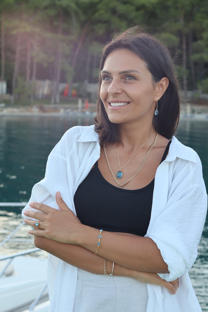 May include: A woman wearing a white shirt, black tank top, and white pants. She is wearing a gold necklace with a blue gemstone pendant, a gold bracelet with blue gemstones, and a gold ring with a blue gemstone. She is smiling and looking to the right.