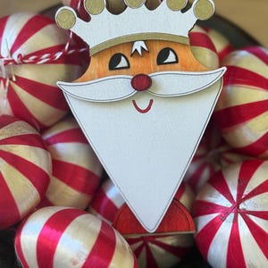 May include: A wooden Christmas ornament in the shape of a king with a white beard, red nose, and gold crown. The ornament is surrounded by red and white striped ornaments, creating a festive holiday scene.