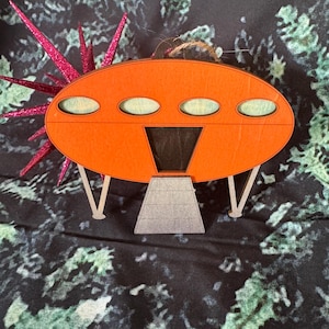 以下が含まれることがあります： オレンジとグレーのウッド製のUFOオーナメント。3つの窓とランディングギアが付いています。