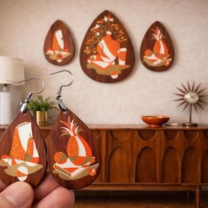 Op de afbeelding: Houten druppelvormige oorbellen met een retro stillevenontwerp van fruit in oranje, wit en goud. De oorbellen hangen voor een interieur in mid-century modern stijl met bijpassende wandkunst en meubels.