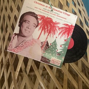 May include: A vintage record album cover featuring a man in a Hawaiian shirt with a Christmas tree and palm trees in the background. The album is titled "Christmas at the Hawaiian Village with Alfred Apaka and the Hawaiian Village Serenaders".