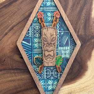Puede incluir: Arte de pared de madera con temática tiki en forma de diamante. El diseño presenta una máscara tiki central, antorchas cruzadas con llamas y hojas estilizadas. El fondo es una tela estampada azul y blanca, enmarcada por un borde de madera.