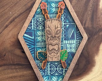 May include: Wooden tiki-themed wall art in a diamond shape. The design features a central tiki mask, crossed torches with flames, and stylized leaves. The background is a blue and white patterned fabric, framed by a wooden border.