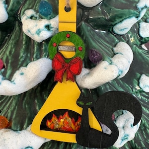 以下が含まれることがあります： 赤いリボンと赤い実のついた緑のリースが付いた、黄色い木製の暖炉の形をしたクリスマスオーナメント。暖炉の前には黒い猫のシルエットがあります。暖炉の中には炎の切り抜きがあります。