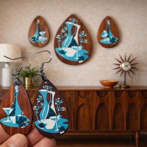 Op de afbeelding: Druppelvormige houten oorbellen met een modern design uit het midden van de eeuw. De oorbellen hebben een blauw-witte illustratie van een vaas, glazen en bloemelementen. De achtergrond omvat bijpassende wandkunst en een houten kast.