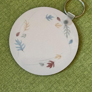 May include: A round white keychain with a brown, yellow, and blue leaf design. The keychain has a silver metal ring.