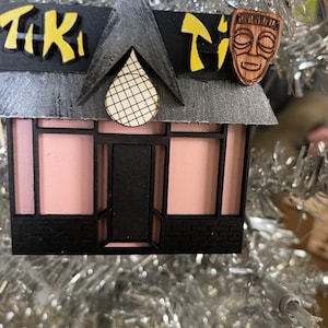 Puede incluir: Adorno navideño de madera de un bar Tiki. El adorno presenta un edificio rosa con adornos negros, un techo gris y las palabras "Tiki Tiki" en letras amarillas. Una máscara Tiki de madera está unida al techo.