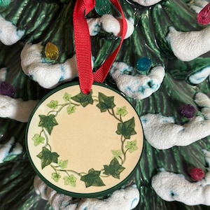 May include: A ceramic Christmas tree with white snow and green leaves. The tree has a red ribbon with a round ornament with a green ivy design.