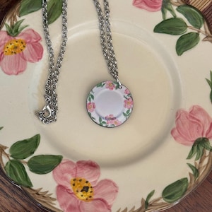 May include: A silver chain necklace with a small, round pendant featuring a white porcelain plate with a pink floral design.