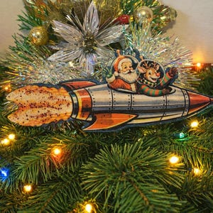 Puede incluir: Adorno navideño con Papá Noel y un reno en un cohete retro. El cohete es plateado y naranja con un escape brillante. El adorno está sobre un fondo de ramas de pino y luces.