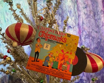 May include: A vintage Christmas tree decorated with ornaments and a vinyl record album cover. The album cover features the text "Christmas with The Chipmunks" and illustrations of the characters. The ornaments include red and gold striped baubles and green and gold spheres.