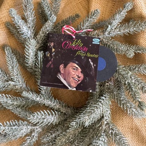 以下が含まれることがあります： フランク・シナトラが写った、ヴィンテージのクリスマスレコードアルバムのオーナメント。タイトルは「A Jolly Christmas from Frank Sinatra」。オーナメントは赤と白のリボンで飾られており、雪が積もった人工の常緑樹の枝で囲まれています。