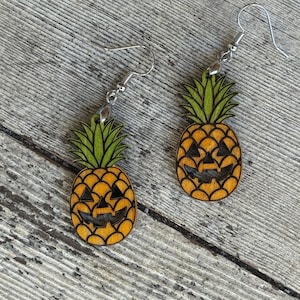 May include: A pair of wooden earrings shaped like jack-o'-lantern pineapples. The earrings feature orange and black carved faces, green leafy tops, and silver-colored ear wires. The earrings are set against a wooden background.