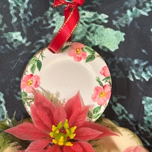 May include: A white ceramic ornament with a red ribbon and a floral design. The ornament features pink roses and green leaves. The ornament is sitting on top of a red poinsettia.
