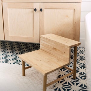 May include: A wooden step stool with two steps, made of light wood. The stool is in front of a bathroom vanity with a white countertop and light wood cabinets. The vanity has a black drawer pull and black round cabinet knobs.
