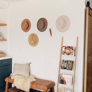 May include: A wooden ladder shelf with five magazines on it. The magazines are titled "Magnolia", "Coastal Living", "Traditional Home", "The New Yorker", and "Food & Wine". The shelf is next to a wooden bench with a white faux fur throw and a tan pillow. There are four hats hanging on the wall behind the bench.