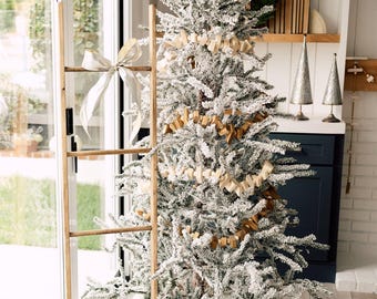 May include: A flocked Christmas tree decorated with a wooden star topper, a wooden garland, and a wooden ladder leaning against the side. The base of the tree is surrounded by gold polka-dot wrapped gifts with silver and gold ribbons.