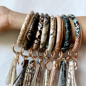 以下が含まれることがあります： 8個のカラフルなヘビ柄キーホルダーのセット。金色のリングとタッセルが付いています。キーホルダーは革製で、さまざまな色とパターンがあります。