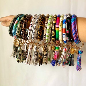 May include: A set of 14 colorful silicone keychains with tassels. Each keychain features a different pattern, including floral, geometric, and animal prints. The keychains are attached to gold-colored rings.