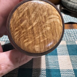 May include: A circular wooden object featuring a light tan center with a distinct grain pattern, surrounded by a dark brown outer ring. The surface has a smooth, polished finish that highlights the natural wood texture and contrasting color tones.