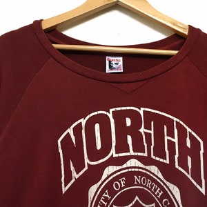 May include: Burgundy red sweatshirt with a white graphic of the word "NORTH" in a vintage style font. The graphic also includes a circular design with the text "University of North Carolina".