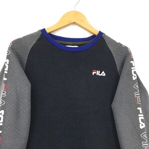 May include: A black and gray long-sleeved sweatshirt with a blue and green trim around the neck. The sweatshirt has the word "FILA" printed on the front in white. The sleeves have the word "FILA" printed in white on a gray background.