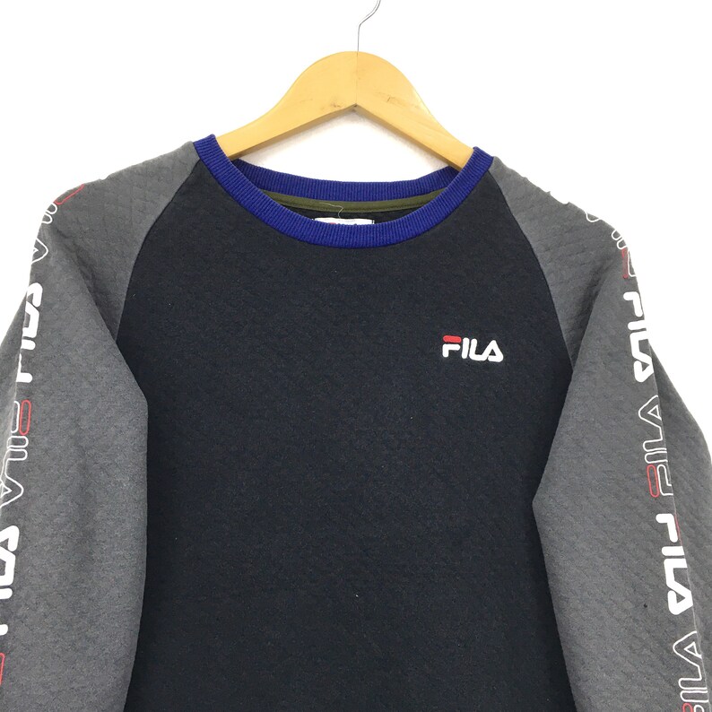 May include: A black and gray long-sleeved sweatshirt with a blue and green trim around the neck. The sweatshirt has the word "FILA" printed on the front in white. The sleeves have the word "FILA" printed in white on a gray background.