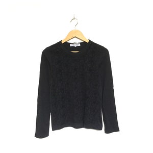 Può includere: Maglia nera a maniche lunghe con motivo floreale sul davanti. La maglia è appesa a una gruccia di legno su sfondo bianco. Il nome del marchio è visibile sul colletto.