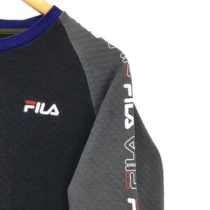 May include: Black and gray long-sleeve sweatshirt with a white Fila logo on the left chest and the word "Fila" repeated down the right sleeve in white with red accents.