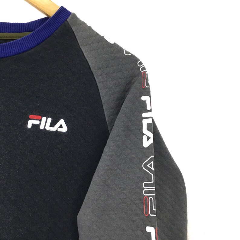 May include: Black and gray long-sleeve sweatshirt with a white Fila logo on the left chest and the word "Fila" repeated down the right sleeve in white with red accents.