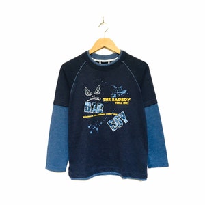 May include: A blue long-sleeve shirt with a graphic print of a cartoon face with the text "The Badboy Since 1997" and "Bad Boy" in yellow lettering.