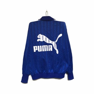 May include: A blue vintage Puma track jacket with a white Puma logo and text on the back. The jacket has a collared neckline and ribbed cuffs and hem. The fabric has a subtle vertical stripe pattern.