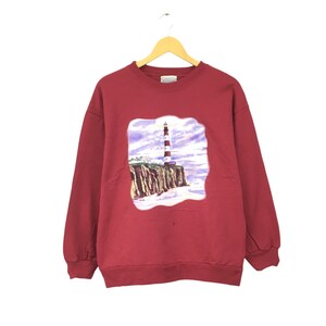 May include: Red long-sleeved sweatshirt with a graphic print of a lighthouse on a cliff. The graphic is in shades of blue, purple, white, and brown.