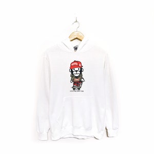 Puede incluir: Sudadera con capucha blanca con un mono de dibujos animados que lleva un gorro rojo y sostiene una patineta. El mono está rodeado de un patrón de camuflaje. El texto "LIVE BY THE STREET CODE" está impreso debajo del mono.
