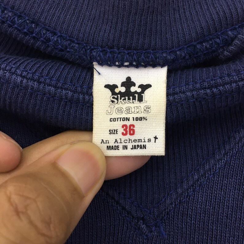 May include: A navy blue cotton t-shirt tag with the text "Skull Jeans" in white, a crown logo, and the text "Cotton 100%", "Size 36", "An Alchemist", and "Made in Japan".
